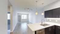 This single-floor space showcases a thoughtfully designed open-concept layout that seamlessly connects the kitchen and living area, creating a sense of spaciousness and flow. The kitchen is appointed with sleek white quartz countertops, dark cabinetry, a stainless steel double-basin sink, and a stainless steel dishwasher. Elegant pendant lights with warm-toned shades hang above the kitchen counter, adding a stylish focal point to the space. A track lighting fixture in the living area provides adjustable, functional illumination throughout. The living space features smooth, light-toned hardwood-style flooring that extends throughout, complementing the crisp white walls and ceilings. Natural light filters in through a large window at the far end of the room, brightening the open interior. A secondary door, likely leading to a utility or entry area, adds convenience to the layout. The overall ambiance is clean, contemporary, and welcoming — ideal for both everyday living and entertaining.