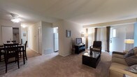 This single-floor residence offers a straightforward and functional layout designed for comfortable everyday living. The living area is generously proportioned, finished with wall-to-wall carpeting in a warm neutral tone that adds a soft, inviting feel underfoot. Large vertical blinds along the exterior wall allow for adjustable natural light, brightening the space and creating an airy atmosphere. The living room provides ample space for seating and entertainment arrangements, with smooth, neutral-toned walls that lend a clean and versatile backdrop. An open sightline connects the living area directly to the kitchen, which is visible in the background and features a full-size refrigerator, contributing to a seamless flow between the two spaces. A bifold closet door is also visible near the entryway, offering convenient built-in storage. The floor plan is efficient and easy to navigate, with a cohesive design throughout. Overall, the space presents a calm, neutral aesthetic with practical features that make it well-suited for a range of living arrangements.