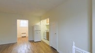 This single-floor space is defined by its warm hardwood floors that extend throughout the main living area, lending a classic character to the interior. The layout is straightforward and functional, with an open main room that receives natural light through a side window, creating a bright and airy atmosphere. A dedicated kitchen is accessible through a doorway from the main living space. The kitchen is outfitted with wood-toned cabinetry, a gas range, and a full-size refrigerator, providing all essential cooking amenities in a compact, efficient arrangement. The neutral, light-painted walls throughout contribute to a clean and understated aesthetic, allowing the natural tones of the flooring to stand out. A secondary door off the main area leads to an additional room or closet space, adding a degree of versatility to the floor plan. A cast-iron radiator is visible along one wall, indicating a traditional heating system. The overall design is utilitarian and straightforward, suited to those who value a no-frills, functional living environment with classic architectural details such as paneled doors and simple trim work that reflect an older but well-maintained construction style.