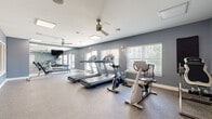 Located at 4011 S Belt Line Rd in Balch Springs, TX, this fitness center is a well-appointed exercise facility designed to meet a variety of workout needs. The single-floor space is finished with durable speckled flooring in the cardio and equipment areas, transitioning to carpet in the open stretching and free-weight zones, providing both functionality and comfort underfoot.

The room is bathed in natural light courtesy of multiple large windows fitted with white blinds, while dual ceiling fans help maintain a comfortable environment throughout workouts. Skylights further brighten the space, contributing to an open and airy atmosphere. The walls are painted in a calm slate blue, creating a clean, modern aesthetic.

Cardio equipment includes multiple treadmills and stationary bikes, positioned to take advantage of the natural light and views to the outdoors. A glass door provides direct access to the exterior, offering views of the surrounding grounds. A wall-mounted television serves as entertainment for cardio sessions.

An adjacent area features a weight bench and additional equipment, with interior windows connecting the fitness center to a neighboring lounge or community room. Posted fitness center rules and clearly marked emergency information ensure a safe and organized environment. Overall, this facility offers a comprehensive and inviting space for residents to pursue their fitness goals.