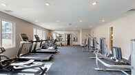 Located at 18183 Wheeler Rd in Westfield, Indiana, this fitness center offers a well-appointed, single-floor exercise room designed to support a wide range of workout routines. The space is finished with neutral-toned walls, recessed lighting throughout, and durable grey carpeting that lends a clean, modern aesthetic.

The exercise room is generously sized and thoughtfully arranged to maximize functionality. Cardio equipment lines one side of the room and includes multiple treadmills, elliptical machines, a stationary upright bike, and a recumbent bike, all positioned to take advantage of natural light streaming in through large windows that offer views of the surrounding community. A wall-mounted flat-screen TV provides entertainment during workouts.

The strength training section features a multi-station Hoist functional trainer as a centerpiece, accompanied by various selectorized weight machines for targeted muscle training, a rowing machine, and a flat utility bench. A rack of dumbbells is also available for free-weight exercises. Full-length mirrors along one wall visually expand the space and allow users to monitor their form.

The room benefits from ample recessed ceiling lighting, ensuring a bright and energizing atmosphere at any time of day. Additional doors provide access to adjacent utility or storage areas, contributing to the overall practicality of this well-equipped fitness facility.