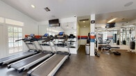 Located at 4801 47th Ave W in Bradenton, Florida, this single-floor fitness center is a comprehensive facility designed to support a full range of workout routines. The space is bright and open, with white walls accented by bold orange and blue horizontal stripes that give the gym an energetic, motivating atmosphere.

The cardio section features a row of treadmills equipped with individual digital displays, allowing users to track their workouts with ease. Complementing these are elliptical trainers, a stationary bike, and a rowing machine, offering a well-rounded selection of low-impact and high-intensity cardio options. A wall-mounted flat-screen television is positioned to keep exercisers entertained during sessions.

The strength training area is equally well-appointed, housing a multi-station cable machine, a weight bench, dumbbells, and a variety of free weights. Full-length mirrors line the walls throughout, enhancing the sense of space and allowing for proper form checks during exercise.

Large French doors and generous windows flood the space with natural light while providing views of the surrounding outdoor area. Ceiling fans help maintain comfortable air circulation. The durable flooring throughout the gym is designed to withstand heavy equipment and high-traffic use. Overall, this fitness center delivers a clean, functional, and well-maintained environment suited for both casual and dedicated fitness enthusiasts.