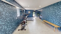 This fitness center is a thoughtfully designed workout facility that combines functionality with a bold, contemporary aesthetic. The most striking design feature is the vivid blue abstract-patterned wallcovering that envelops the space, creating an energetic and motivating environment reinforced by an inspirational "you go" wall phrase. Light-toned tile flooring runs throughout, providing a clean contrast to the deep blue walls, while recessed ceiling lighting ensures the space is consistently well-lit.

The facility is comprehensively equipped for a full-body workout. Cardio equipment includes multiple treadmills, an elliptical trainer, and a stationary bike, all positioned to make use of the natural light streaming in through windows along one wall. A dedicated free weights area features a complete rack of dumbbells, accompanied by a weight bench for strength training. A rowing machine rounds out the equipment selection, offering users a diverse range of exercise options.

Additional amenities include a water dispenser, a wall-mounted HVAC unit for climate control, and flat-screen televisions mounted on the walls for entertainment during workouts. A wooden bench provides a convenient space for stretching or changing footwear. A private restroom or changing area is accessible via a separate door. The facility is generously sized, allowing ample open floor space for movement and exercise between equipment stations.