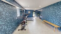 This fitness center is a thoughtfully designed workout facility that combines functionality with a bold, contemporary aesthetic. The most striking design feature is the vivid blue abstract-patterned wallcovering that envelops the space, creating an energetic and motivating environment reinforced by an inspirational "you go" wall phrase. Light-toned tile flooring runs throughout, providing a clean contrast to the deep blue walls, while recessed ceiling lighting ensures the space is consistently well-lit.

The facility is comprehensively equipped for a full-body workout. Cardio equipment includes multiple treadmills, an elliptical trainer, and a stationary bike, all positioned to make use of the natural light streaming in through windows along one wall. A dedicated free weights area features a complete rack of dumbbells, accompanied by a weight bench for strength training. A rowing machine rounds out the equipment selection, offering users a diverse range of exercise options.

Additional amenities include a water dispenser, a wall-mounted HVAC unit for climate control, and flat-screen televisions mounted on the walls for entertainment during workouts. A wooden bench provides a convenient space for stretching or changing footwear. A private restroom or changing area is accessible via a separate door. The facility is generously sized, allowing ample open floor space for movement and exercise between equipment stations.