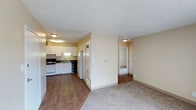 This single-floor unit is thoughtfully laid out to maximize functionality across its living spaces. The open-concept design connects the kitchen directly to the living area, creating a cohesive and practical flow throughout. Warm beige walls and textured ceilings are consistent throughout the unit, lending a neutral, move-in-ready aesthetic.

The kitchen is well-equipped with white cabinetry offering ample upper and lower storage, dark countertops, a gas range, dishwasher, refrigerator, and a single-basin sink. Hardwood-style flooring runs through the kitchen, complementing the bright flush-mount lighting overhead.

The living area transitions to plush carpeting, providing a comfortable and quiet space. A large window fitted with vertical blinds and a wall-mounted air conditioning unit ensures both natural light and climate control. An additional carpeted room, accessible from the living area, offers flexible use as a bedroom or secondary living space, also finished with neutral walls and clean lines.

The bathroom features a full-size soaking tub with a shower and curtain rod, paired with an over-toilet cabinet for convenient storage. Clean white fixtures and a simple, functional design define the space.