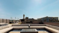 This penthouse-level space serves as the gateway to a stunning rooftop terrace, combining modern interior design with seamless outdoor access. The interior is defined by expansive floor-to-ceiling windows framed in sleek black steel, flooding the space with abundant natural light and offering unobstructed views of the surrounding cityscape. Wood-look tile flooring in warm, natural tones adds a sophisticated yet welcoming aesthetic throughout.

A bold black glass door provides direct access to the rooftop terrace, where a generously sized outdoor lounge area awaits. The terrace is thoughtfully furnished with large sectional sofas in neutral upholstery, paired with low-profile coffee tables including a fire pit table, all arranged to maximize comfort and the sweeping urban views. Horizontal slatted privacy screens define the perimeter, adding a contemporary architectural detail while maintaining an open-air ambiance.

The rooftop deck is finished with light-toned pavers that complement the clean, modern aesthetic of the overall space. Tiered levels on the terrace offer varied seating arrangements, with lush greenery and planters adding natural accents against the open sky. The combination of the bright, airy interior and the expansive outdoor terrace makes this a distinctive, versatile space ideal for gathering and entertaining at elevation.