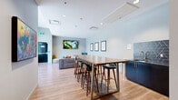 Located at 233 Kennedy St in Winnipeg, MB, this single-floor communal living space is designed with a contemporary aesthetic and a welcoming, functional layout. Light hardwood flooring runs throughout, complementing the soft grey and white tones of the walls and creating a bright, airy atmosphere.

The space is divided into two distinct zones that flow seamlessly into one another. The lounge area features a large sectional sofa accented with colorful throw pillows, a woven pouf ottoman, a small coffee table, and a cozy accent armchair — all oriented around a wall-mounted flat-screen TV, making it an inviting spot for relaxation or socializing. A second wall-mounted TV is positioned nearby, further enhancing the entertainment options.

Toward the other end of the room, a sturdy bar-height communal table with wood-and-metal construction is surrounded by stylish bar stools, offering a casual space for dining, working, or gathering. Adjacent to the table, a sleek dark-tiled wet bar area provides additional utility.

Large windows along one wall flood the space with natural light and offer views of the surrounding Winnipeg streetscape. Recessed ceiling lighting supplements the natural light throughout. Framed artwork and urban photography panels add personality and a polished finish to this thoughtfully designed shared space.