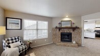This single-floor home is designed with a clean, neutral aesthetic that creates a warm and functional living environment. The living room serves as a central gathering space, anchored by a striking floor-to-ceiling stone fireplace with a dark wood mantel, complemented by plush carpeting and multiple windows that allow natural light to fill the room. A front entry area adjacent to the living space features additional windows, enhancing the bright and open feel.

The kitchen is outfitted with white cabinetry, grey speckled countertops, a double-basin stainless steel sink, and dark hardwood-style flooring. Black appliances, including a range, dishwasher, and refrigerator, provide a bold contrast to the light cabinetry. The layout offers efficient counter space and storage throughout.

The bedrooms are generously sized, carpeted, and painted in soft grey tones. One bedroom features a sliding-door closet and a window with natural light, while another connects to a bathroom area with a dedicated vanity station — complete with a speckled countertop, built-in drawers, and a well-lit mirror. The overall floor plan, as seen in the dollhouse view, offers a practical and well-organized layout suited to comfortable everyday living.