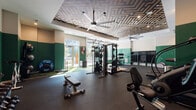 This modern fitness center is thoughtfully designed to support a wide range of workout routines. The space is anchored by a bold aesthetic, highlighted by a striking geometric-patterned ceiling with large industrial ceiling fans that provide both visual impact and ventilation throughout the room. Recessed lighting complements the natural light streaming in through oversized windows and glass doors, which open to an outdoor area and provide views of the surrounding environment.

The flooring transitions between durable rubber matting and artificial turf, delineating functional zones within the gym. The functional training area features cable machines, a power rack, and a dedicated turf zone outfitted with kettlebells, medicine balls, and battle ropes — ideal for dynamic and high-intensity workouts. A cardio section lines one wall with a row of treadmills and upright bikes, accompanied by a wall-mounted TV for entertainment. Additional cardio equipment includes a stair climber and a stationary recumbent bike.

Strength training is further supported by a dumbbell rack and flat utility benches. Green accent walls add a vibrant, energetic tone to the space, while open shelving and storage cubbies keep equipment organized. A frosted glass-paneled room provides a semi-private area within the fitness center. The overall design is clean, modern, and purpose-built for performance.