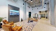 Located at 628 Ellicott St in Buffalo, NY, this single-floor lobby space offers a striking industrial-chic aesthetic that blends raw architectural elements with contemporary design. Soaring ceilings with exposed ductwork, structural concrete columns, and pendant lighting create an open, loft-like atmosphere throughout the generously sized floor plan.

The main lounge area anchors the space with a vibrant, multi-colored terrazzo-style area rug, comfortable tan channel-tufted seating, and a sleek round coffee table arrangement. A large-format display screen mounted on a dark tile feature wall adds a bold focal point. Polished concrete floors extend throughout, lending a clean, cohesive look that complements the industrial architecture.

Toward the front of the building, floor-to-ceiling windows flood a secondary lounge nook with abundant natural light and offer views of the surrounding Buffalo cityscape. This area includes a built-in credenza with cabinetry, a beverage station, and a banquette seating arrangement — ideal for casual gatherings. A dedicated mailroom, finished in deep teal with wall-to-wall black parcel lockers and individual mailboxes, provides practical resident amenities. Private rooms with glass-panel doors line the corridor, adding functional versatility to this well-appointed building lobby.