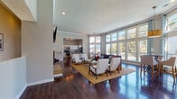 This single-floor clubhouse is designed with an open, flowing layout that seamlessly connects its key areas, making it an ideal communal gathering space. Rich, dark hardwood flooring runs throughout, complemented by crisp white trim, crown molding, and a neutral color palette that lends the space a clean, polished aesthetic.

The main lounge area is anchored by a tiled fireplace with a white mantel, above which a wall-mounted flat-screen television is installed. Generous seating, including a dark upholstered sofa and mid-century-style armchairs arranged on a natural fiber area rug, creates an inviting atmosphere for residents to relax and socialize. Soaring ceilings with recessed lighting further enhance the sense of openness throughout.

Floor-to-ceiling windows along one wall flood the interior with abundant natural light while providing direct views of the outdoor pool and surrounding courtyard. A dedicated dining nook, furnished with a high-top table and chairs beneath a woven pendant light, takes full advantage of these views, with patterned draping framing the expansive glazing.

The full kitchen is equipped with stainless steel appliances — including a range, microwave, and refrigerator — white shaker cabinetry, granite countertops, and a breakfast bar peninsula, providing everything needed to support events and gatherings. The overall ambiance of the clubhouse is bright, modern, and community-oriented.