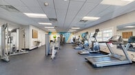 Located at 150-A Willowdale Dr in Frederick, Maryland, this well-appointed fitness center offers a spacious, single-floor layout designed to accommodate a comprehensive workout experience. The open floor plan is finished with durable rubberized flooring throughout, providing traction and comfort during exercise. A drop-panel ceiling with recessed fluorescent lighting ensures the space is consistently and brightly illuminated, while multiple windows along the perimeter walls introduce natural light and views of the surrounding outdoors.

The main workout area is equipped with an extensive array of cardio machines, including treadmills, ellipticals, and a rowing machine, all arranged to maximize accessibility and traffic flow. A dedicated strength training section features a multi-function cable machine and various resistance equipment, catering to a range of fitness routines. A vibrant, large-scale abstract mural serves as a dynamic focal point along one wall, adding energy and visual interest to the environment.

An entry area near the front door provides organized storage cubbies and a waste receptacle, offering a practical transition space for users. Additional private rooms accessible via interior doors offer further utility. The overall ambiance is clean, functional, and motivating, making this fitness center a well-equipped facility for residents of the Willowdale community in Frederick, MD.