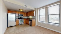Located at 367 E Broad St in the heart of Columbus, Ohio, this single-floor unit offers a well-designed layout that balances functional living with urban convenience. The kitchen is a standout feature, outfitted with warm honey-toned wood cabinetry, dark countertops, and a full suite of stainless steel appliances including a refrigerator, electric range, built-in microwave, and dishwasher. Tile flooring in the kitchen transitions seamlessly into plush carpeting throughout the remainder of the unit. The living area is generously proportioned, bathed in natural light through multiple large windows that frame city views of the Columbus skyline. Neutral beige walls and clean white ceilings create a bright, airy atmosphere throughout. A carpeted entryway corridor leads into the main living space, providing a welcoming arrival experience. The bathroom is finished with tile flooring, a full-size tub and shower surround, and wood-toned cabinetry. Throughout the unit, consistent neutral tones, ample natural light, and practical finishes combine to create a comfortable and functional urban living environment in a prime downtown Columbus location.