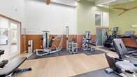 The fitness center at 100 Buckingham Dr in Santa Clara, California, is a thoughtfully designed gym space that offers a comprehensive workout environment. The facility is characterized by striking vaulted ceilings with white-painted wood paneling, accented by sleek linear lighting and a modern ceiling fan that enhances airflow and comfort throughout.

The interior features a warm, inviting aesthetic with sage green upper walls, a warm wood-toned wainscoting detail, and light hardwood-style flooring complemented by protective rubber fitness mats beneath the equipment. Large arched windows and glass doors flood the space with abundant natural light and offer views of the surrounding landscaping, creating an open and energizing atmosphere.

The gym is well-equipped with a strong selection of Cybex cardio machines, including treadmills, upright and recumbent bikes, and a StairMaster, as well as a variety of Cybex strength machines for targeted muscle training. A dedicated free weights area features a full dumbbell rack with a flat bench, and additional functional fitness equipment rounds out the offerings. The layout is organized to allow for comfortable movement between stations. An adjacent entry area connects the gym to the broader community, making this fitness center a convenient and polished amenity for residents of the 100 Buckingham Dr community.