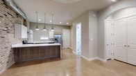 This contemporary two-floor space showcases a thoughtfully designed interior with a clean, modern aesthetic throughout. The open-concept layout on the main level seamlessly connects the kitchen and living areas, creating a spacious and functional environment. Polished concrete floors reflect natural and recessed lighting, lending the space a bright, refined character.

The kitchen is a standout feature, anchored by a substantial island with a rich wood-toned base and crisp white countertop surface. Three industrial-style pendant lights hang above the island, adding both task lighting and a stylish focal point. Cabinetry combines white upper cabinets with warm wood-toned lower units, complementing the neutral gray wall tones. A stainless steel French-door refrigerator with a built-in water and ice dispenser adds a premium touch, while a window above the sink area allows natural light to filter in.

The walls are finished in a soft, warm gray palette that pairs harmoniously with the white baseboards and trim throughout. A frosted glass door panel introduces an elegant architectural detail, diffusing light while maintaining privacy. Recessed ceiling lights are distributed evenly, ensuring consistent illumination across the open floor plan. The overall ambiance is modern, clean, and welcoming, with a cohesive design language carried across both levels of the space.