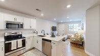This single-floor, 2-bedroom, 1-bathroom residence offers a thoughtfully designed open-concept layout that maximizes its compact footprint. The kitchen and living area flow seamlessly together, anchored by a peninsula with quartz countertops that doubles as a breakfast bar with barstool seating. The kitchen is well-appointed with white shaker cabinetry featuring matte black hardware, stainless steel appliances including a refrigerator, electric range, microwave, and dishwasher, complemented by a deep undermount sink with a matte black faucet. Light wood-look flooring runs throughout the main living area, enhancing the bright, airy atmosphere created by recessed lighting and a large window that draws in natural light.

The living room provides a comfortable gathering space with room for a sectional sofa and entertainment setup. Both bedrooms feature neutral carpet, windows with exterior views, and sufficient space for a queen-sized bed and furnishings. The primary bedroom is a well-proportioned private retreat. The shared bathroom is finished in a clean, modern style with white subway tile surrounding a tub-shower combo accented by matte black sliding glass doors and fixtures, a white vanity, and wood-look flooring. A dedicated closet completes the unit's storage offerings.