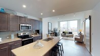 This contemporary single-floor residence showcases a thoughtfully designed open-concept layout that seamlessly blends the kitchen, dining, and living areas into one cohesive, flowing space. The kitchen is a standout feature, outfitted with rich dark-toned cabinetry, stainless steel appliances including a built-in microwave and range, and a generous island with a light quartz countertop and an undermount sink. The island doubles as a breakfast bar with seating, making it ideal for casual dining and entertaining. Recessed lighting throughout the ceiling adds a clean, modern ambiance while ensuring the space is well-lit. The living area extends naturally from the kitchen, bathed in an abundance of natural light streaming through expansive floor-to-ceiling windows. These windows frame sweeping views of a vibrant urban skyline, creating a dramatic and ever-present connection to the city beyond. The flooring is consistent throughout, reinforcing the open and airy feel of the space. Neutral tones and modern finishes are carried throughout, lending the residence a refined, contemporary aesthetic that is both functional and visually striking.