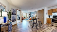 This single-floor residence offers a thoughtfully designed layout that balances comfort and functionality across its key living areas. The open-concept living and dining space is anchored by durable wood-look flooring that flows throughout the unit, creating a cohesive and inviting atmosphere. The living room features a striking bay window arrangement that draws in abundant natural light, complemented by a textured ceiling and a soothing blue accent wall that adds depth and character to the space. A high-top dining table with bar stools provides a casual dining area that transitions seamlessly into the kitchen. The kitchen is equipped with warm honey-toned cabinetry, stainless steel appliances including a range, microwave, and refrigerator, and a functional L-shaped layout that maximizes workspace and storage. The primary bedroom is a comfortable retreat, featuring a large window overlooking outdoor greenery, carpeted flooring, and ample room for furnishings. A second bedroom offers flexible use, currently configured as a children's room with playful décor and built-in bunk bed-style sleeping arrangements. The bathroom features a glass-enclosed shower, wood-look flooring, and clean white walls. Throughout the residence, warm wood-toned door and window trim provide a consistent, welcoming design element.