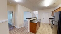 This single-story home in Greenwood, Indiana offers a functional and well-organized layout across three bedrooms and two bathrooms. The open-concept design connects the kitchen, dining, and living areas, creating a cohesive central living space. An arched passageway adds a subtle architectural detail, lending character to the otherwise clean, neutral interior.

The kitchen is equipped with warm-toned wood cabinetry, dark countertops, black appliances including a range, microwave, and refrigerator, and a distinctive wine rack cabinet detail. A peninsula with matching countertops provides additional workspace and separates the kitchen from the living and dining areas. Light ceramic tile flooring runs throughout the kitchen.

The living and dining areas feature light wood-style flooring and neutral walls, with crown molding adding a refined touch. A ceiling fan with a window view — which overlooks what appears to be a water feature — enhances the primary living area. Bedrooms are carpeted and bright, with windows allowing natural light. One bedroom features a double closet with arched panel doors. Both bathrooms include wood vanities with dark countertops and light flooring, maintaining a consistent aesthetic throughout the home.