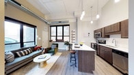 This single-floor space showcases a thoughtfully designed open-concept layout that seamlessly integrates the living and kitchen areas into one cohesive environment. Soaring ceilings with exposed ductwork lend an industrial character to the space, while light-toned walls and wide-plank hardwood-style flooring create a warm, contemporary aesthetic throughout.

The kitchen is a standout feature, outfitted with dark espresso cabinetry, stainless steel appliances — including a range, microwave, and dishwasher — and a generous island topped with a crisp white countertop. Pendant lighting above the island adds both function and style, and bar seating makes the space ideal for casual dining or entertaining.

The adjacent living area is bathed in natural light, courtesy of large black-framed windows that frame views of the surrounding urban landscape. The space accommodates a comfortable seating arrangement with a sofa, armchairs, and a round marble-top coffee table, creating an inviting atmosphere for relaxation.

Throughout the space, the combination of industrial architectural details, modern finishes, and abundant natural light results in a balanced, urban-contemporary ambiance that is both functional and visually engaging.