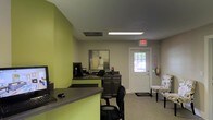 This single-floor commercial space is designed with functionality and professionalism in mind. The reception area serves as the focal point upon entry, featuring bold lime-green accent walls that create a vibrant, energetic atmosphere against neutral gray tones throughout the rest of the space. The front desk area is thoughtfully equipped with computer workstations, providing an efficient workspace for staff. Overhead, recessed fluorescent lighting ensures consistent, well-distributed illumination throughout the area. The flooring features neutral carpeting that complements the overall color scheme and adds a sense of warmth to the space. A white paneled entry door with a sidelight window allows natural light to filter into the reception area, enhancing the brightness of the interior. A seating area near the entrance offers a comfortable waiting space for visitors, furnished with a patterned accent chair. Decorative artwork on the wall adds a polished, professional touch to the environment. Safety features, including an exit sign and fire extinguisher, are visibly integrated into the space. The layout is open and accessible, making it well-suited for a variety of professional or client-facing business operations.