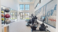 This contemporary fitness center is designed to inspire and energize, offering a thoughtfully equipped workout space bathed in abundant natural light. The room features soaring double-height ceilings with clerestory windows and large floor-to-ceiling glass panels that frame views of the surrounding outdoor grounds, creating an open, airy atmosphere throughout the space.

The cardio zone is outfitted with a lineup of True-brand treadmills, ellipticals, and step machines, all positioned to take advantage of the natural light and outdoor views. A large full-wall mirror along one side of the room amplifies the sense of space and supports functional training. Complementing the cardio equipment is a well-stocked free weights and accessories area, featuring a multi-tiered rack of stability balls in varying sizes and medicine balls for functional fitness training.

A standout design element is the large-scale motivational mural rendered in bold teal brushstroke lettering against a textured gray backdrop, anchoring the main wall and adding a strong visual identity to the space. Neutral-toned carpet flooring, recessed lighting, and a ceiling fan round out the clean, modern interior. The overall ambiance is bright, welcoming, and purpose-built for an effective fitness experience.