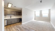 This single-floor unit is positioned at a notable height, as evidenced by the expansive panoramic views visible through its numerous windows. The open-concept layout connects a well-appointed kitchen to a spacious living area, creating a cohesive and functional flow throughout the space. The kitchen features gray-toned cabinetry with gold hardware, white countertops, stainless steel appliances including a dishwasher and range, and a deep undermount sink with a matte black faucet, all set over wood-look vinyl plank flooring. The living area transitions seamlessly from hard flooring to plush carpet, adding warmth underfoot. Multiple bedrooms are finished with neutral carpet and large windows fitted with roller shades, each capturing broad city skyline and waterway views that fill the rooms with abundant natural light. A bathroom includes a full-size soaking tub with a clean white surround. Throughout the unit, neutral warm-gray walls, crisp white baseboards, and consistent finishes contribute to a clean, modern aesthetic. The elevated vantage point is a defining characteristic, making natural light and outdoor scenery a prominent feature of nearly every room.