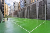 This facility features a full-size outdoor tennis court finished in a green hard-court surface, with crisp white line markings and a central net in place. The court is set within a sheltered urban courtyard, enclosed by a distinctive curved brick wall and tall safety netting supported by steel posts, providing both containment and a striking architectural backdrop. Surrounding multi-story buildings with brick facades rise on all sides, framing an open sky above and creating a well-defined, contained playing environment. Courtside seating benches offer a practical amenity for players and spectators alike.

Access to the interior is provided through a secure, keycard-controlled entry door featuring a full-length brushed stainless steel pull handle and a modern electronic access panel. The interior corridor is clean and functional in design, with smooth white walls, recessed ceiling lighting, and patterned grey carpet tiles that run the length of the hallway. Emergency exit signage is clearly visible, reflecting a well-maintained and safety-conscious facility. The overall ambiance is modern and purposeful, combining robust outdoor athletic amenities with a secure, professionally finished interior passage.