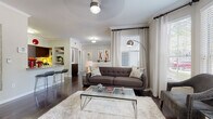 This single-story residence showcases a thoughtfully designed open-concept layout that seamlessly connects the living, dining, and kitchen areas. Crown molding and rich dark hardwood flooring run throughout the main living spaces, contributing to a polished, cohesive aesthetic.

The living room is bathed in natural light from multiple windows dressed with sheer white curtains and blinds, including a distinctive bay window arrangement that expands the sense of space. The kitchen features dark espresso cabinetry, granite countertops, stainless steel appliances including a refrigerator, range, and built-in microwave, and an island with a sink that overlooks the living area. A breakfast bar with seating provides an additional casual dining option.

The dedicated dining area accommodates a glass-top table and sits adjacent to a door offering outdoor views and additional natural light. The bedrooms are carpeted for comfort, each offering a calm, neutral atmosphere. One bedroom features a tufted upholstered headboard and warm accent lighting, while another presents a serene, similarly styled retreat.

The bathroom includes a dark-stained vanity with granite countertops, a framed mirror, tile flooring, and built-in shelving, completing this well-finished residence.