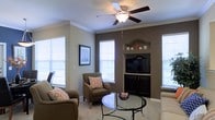 This single-floor residence offers a thoughtfully designed living space that balances comfort and classic style. The living room serves as the heart of the home, showcasing crown molding details along the ceiling and a neutral color palette with a warm accent wall that adds depth and character to the room. A built-in media niche with display shelving above provides a functional and stylish focal point, ideal for a television and decorative accents. The space is finished with plush carpeting underfoot, contributing to a cozy and inviting atmosphere. Multiple windows dressed with white horizontal blinds flood the room with natural light, creating an open and airy feel. A ceiling fan with a light fixture is centrally positioned, ensuring year-round comfort and additional illumination. The living area is generously proportioned, comfortably accommodating a full seating arrangement including upholstered armchairs and a glass-top coffee table. The overall ambiance is warm and welcoming, reflecting a well-maintained interior that blends practical features with tasteful design elements throughout.