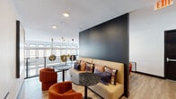 Located on Polaris Point Loop in Colorado Springs, CO, this thoughtfully designed communal lounge space combines modern aesthetics with a warm, inviting atmosphere. The single-floor layout centers around a striking dark textured accent wall that anchors the seating area, where a generous camel-toned banquette sofa is dressed with an eclectic mix of patterned throw pillows in rich purples, pinks, and earth tones. Flanking the seating are terrazzo-topped bistro tables paired with plush, rust-orange accent chairs, creating intimate conversation nooks throughout the space. Warm-toned, light wood plank flooring runs throughout, lending continuity and a natural warmth to the interior. A cluster of globe pendant lights in brushed gold hangs near an open-railing overlook, adding a sculptural focal point and drawing the eye toward the generous windows that allow natural light to pour in. Exposed metallic ductwork lends an industrial edge to the corridor, which is lined with dark-framed doors and features slatted wood wall panels for visual texture. A large-scale abstract artwork adds a creative, contemporary accent to the gallery-style wall near the overlook. The overall ambiance is modern and curated, balancing bold design choices with comfortable, functional furnishings suited for communal use.