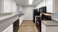 Located at 584 E Barham Dr in San Marcos, CA, this single-floor unit offers a well-appointed, functional layout with modern finishes throughout. The galley-style kitchen is equipped with white shaker cabinetry, gray countertops, a stainless steel sink, and a full suite of black appliances including a range, microwave, and dishwasher. Wood-style plank flooring runs throughout the unit, lending warmth and visual continuity from room to room. The open living area is bright and spacious, featuring neutral gray walls, a textured ceiling, and a sliding glass door fitted with vertical blinds that provides access to an outdoor balcony or patio and allows natural light to fill the room. A wall-mounted air conditioning unit services the living space. The kitchen connects to the living area via a pass-through opening, creating an inclusive, connected feel. The bathroom features a white shaker-style vanity with gray countertop, a large mirror with overhead lighting, wood-style flooring, and a shower-tub combination. Additional interior doors suggest dedicated storage or closet space. The overall aesthetic is clean, contemporary, and move-in ready, reflecting a cohesive modern update throughout the unit.