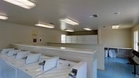 This single-floor communal laundry facility is designed for efficiency and ease of use. The room is laid out with a clear division between washing and drying areas, featuring a row of Whirlpool top-load washing machines positioned along the front and a bank of white dryers lined up along the back wall. A wide, flat countertop surface running between the two sections provides ample space for folding laundry or staging items. The room is brightly illuminated by multiple overhead fluorescent light fixtures evenly spaced across the white ceiling, ensuring a well-lit environment throughout. Ventilation is provided via ceiling-mounted HVAC vents. The walls are finished in a neutral, light tone, contributing to a clean and functional atmosphere. A window at the back of the room allows for some natural light to supplement the artificial lighting. Informational notices and signage are posted on the walls, offering guidance for users of the facility. The flooring is a durable, easy-to-clean surface appropriate for a high-traffic laundry environment. Overall, the space is practical, orderly, and well-maintained, making it a reliable shared laundry resource.