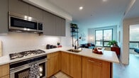 This contemporary single-floor residence showcases a thoughtfully designed open-concept layout that seamlessly connects the kitchen, dining, and living areas. The kitchen is a standout feature, outfitted with warm wood lower cabinetry contrasted by matte gray upper cabinets, white quartz countertops, stainless steel appliances including a gas range with oven and built-in microwave, and an undermount sink. A peninsula with bar seating provides an ideal casual dining space while maintaining visual openness to the living area. The living room is bathed in natural light from large, dark-framed windows offering urban treetop views, creating a bright and inviting atmosphere. Light wood-tone flooring flows throughout the main living spaces, enhancing the sense of continuity and warmth. The bedroom is generously sized with expansive windows framing outdoor views, allowing abundant natural light to fill the room. It features light hardwood flooring and wall-mounted lighting for a clean, modern aesthetic. The bathroom is finished with a floating wood vanity, marble-look quartz countertop, an undermount sink, and a large mirror, reflecting a refined and minimal design sensibility. Overall, the space delivers a cohesive modern aesthetic with quality finishes and a functional, open feel.