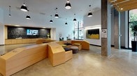 This thoughtfully designed lobby makes a strong first impression through its blend of contemporary architecture and curated artistic elements. At its core is a distinctive, freestanding light wood seating structure that winds organically through the space, offering built-in bench seating accented with navy and green cushions alongside small round tables. Black dome pendant lights hang at varying heights throughout, providing focused illumination across the polished concrete floors.

One section of the lobby transitions into a more intimate area distinguished by an exposed wood-slat ceiling with track lighting, a colorful mosaic-tiled floor, and a large-scale mural that anchors the wall with bold, urban-inspired imagery. Nearby, deep blue open shelving and concrete columns add industrial texture and contrast to the warm wood tones.

Floor-to-ceiling glazing at the entrance frames views of the surrounding streetscape, welcoming abundant natural light and a strong visual connection to the outdoors. A media wall featuring a mounted display sits behind a reception-style counter clad in patterned black tile. Curated artwork, sculptural branches, and potted plants are placed throughout, contributing to a vibrant, gallery-like atmosphere that balances functionality with creative energy.