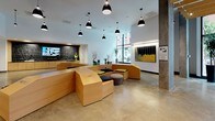This thoughtfully designed lobby makes a strong first impression through its blend of contemporary architecture and curated artistic elements. At its core is a distinctive, freestanding light wood seating structure that winds organically through the space, offering built-in bench seating accented with navy and green cushions alongside small round tables. Black dome pendant lights hang at varying heights throughout, providing focused illumination across the polished concrete floors.

One section of the lobby transitions into a more intimate area distinguished by an exposed wood-slat ceiling with track lighting, a colorful mosaic-tiled floor, and a large-scale mural that anchors the wall with bold, urban-inspired imagery. Nearby, deep blue open shelving and concrete columns add industrial texture and contrast to the warm wood tones.

Floor-to-ceiling glazing at the entrance frames views of the surrounding streetscape, welcoming abundant natural light and a strong visual connection to the outdoors. A media wall featuring a mounted display sits behind a reception-style counter clad in patterned black tile. Curated artwork, sculptural branches, and potted plants are placed throughout, contributing to a vibrant, gallery-like atmosphere that balances functionality with creative energy.