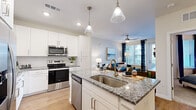 This single-floor residence is designed with a modern, open-concept layout that seamlessly connects the kitchen and living areas. The kitchen is a standout feature, equipped with stainless steel appliances including a range, microwave, dishwasher, and refrigerator. Crisp white shaker-style cabinetry extends to the ceiling, complemented by a white subway tile backsplash and granite countertops with subtle gray and blue tones. A central island with seating provides additional prep space and casual dining, illuminated by elegant pendant lights. Light wood-tone flooring runs throughout the main living areas, enhancing the bright, airy atmosphere.

The living area offers natural light through large windows dressed with patterned curtains, and features a ceiling fan overhead. A door with sidelight provides additional outdoor views. The bedroom is carpeted and includes a ceiling fan and window with exterior views. The bathroom presents a glass-enclosed walk-in shower with tile surround and built-in shelving, with direct access to a walk-in closet. Throughout the space, neutral wall tones and recessed lighting create a clean, cohesive aesthetic.