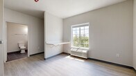 This efficiently designed single-floor studio unit offers a clean, modern aesthetic throughout, making excellent use of its compact footprint. The main living area features light-toned luxury vinyl plank flooring, neutral gray walls, and a large grid-pane window that fills the room with natural light and frames views of surrounding greenery. A wall-mounted fold-down shelf provides a practical workspace or dining surface, while a built-in PTAC unit ensures year-round climate comfort. The kitchen is outfitted with sleek, flat-front white cabinetry, a stainless steel refrigerator, a single-basin sink with a brushed nickel faucet, and light countertops — all arranged in a streamlined galley configuration. An in-unit washer is conveniently tucked beside the kitchen area, adding a valuable practical amenity. The bathroom features dark slate-style tile flooring, a modern vanity with a white countertop, brushed nickel fixtures, and a track lighting bar above the mirror, creating a well-lit and functional space. The unit's consistent use of light tones, contemporary finishes, and thoughtful built-in features contribute to a bright, clean, and highly functional living environment.