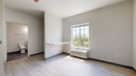 This efficiently designed single-floor studio unit offers a clean, modern aesthetic throughout, making excellent use of its compact footprint. The main living area features light-toned luxury vinyl plank flooring, neutral gray walls, and a large grid-pane window that fills the room with natural light and frames views of surrounding greenery. A wall-mounted fold-down shelf provides a practical workspace or dining surface, while a built-in PTAC unit ensures year-round climate comfort. The kitchen is outfitted with sleek, flat-front white cabinetry, a stainless steel refrigerator, a single-basin sink with a brushed nickel faucet, and light countertops — all arranged in a streamlined galley configuration. An in-unit washer is conveniently tucked beside the kitchen area, adding a valuable practical amenity. The bathroom features dark slate-style tile flooring, a modern vanity with a white countertop, brushed nickel fixtures, and a track lighting bar above the mirror, creating a well-lit and functional space. The unit's consistent use of light tones, contemporary finishes, and thoughtful built-in features contribute to a bright, clean, and highly functional living environment.