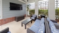 This outdoor amenity space is designed for relaxation and entertainment, centered around a large, sparkling swimming pool surrounded by a generous concrete deck. The pool deck is furnished with multiple lounge chairs and shaded dining areas with umbrellas, providing ample space for sunbathing and casual gatherings.

Adjacent to the pool is a covered outdoor lounge area supported by classic white columns with brick accents at the base. This sheltered space is equipped with ceiling fans to keep the area comfortable, and a large wall-mounted flat-screen television serves as a focal point for outdoor entertainment. The seating area features dark wicker furniture with blue and white striped cushions, including a sofa, armchairs, and a glass-topped coffee table, creating an inviting and cohesive aesthetic.

Decorative metal trellis panels line the perimeter of the covered lounge, allowing natural light and views of the surrounding landscaping to filter through while adding an architectural element to the space. The overall ambiance is that of a well-appointed resort-style retreat, offering both shaded comfort and open-air lounging directly at the water's edge. The multi-story residential buildings visible in the background provide a sense of community scale and context for this shared outdoor living area.