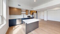 Located on South Temple Street in Salt Lake City, this thoughtfully designed one-bedroom, one-bathroom residence offers a clean, modern aesthetic across a well-appointed single floor. The open-concept kitchen and living area serves as the heart of the home, showcasing light wood-tone flooring, recessed lighting, and an abundance of natural light from large windows. The kitchen is a standout feature, equipped with stainless steel appliances including a range, dishwasher, and refrigerator, complemented by two-tone cabinetry — navy lower cabinets paired with warm wood upper cabinets and open shelving. A sleek center island with a white quartz countertop and a classic white subway tile backsplash complete the contemporary look. The bedroom offers plush carpeting, a ceiling fan, and a window with views of the surrounding cityscape. A sliding barn door leads to an additional space with hardwood-style flooring. The walk-in closet provides generous storage with wrap-around shelving and hanging rods. The bathroom features light wood-tone flooring, a warm wood vanity with a white countertop, and a frameless glass-enclosed walk-in shower with white tile surround and recessed lighting, creating a clean, spa-like atmosphere.