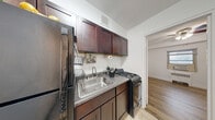 This single-floor unit offers a thoughtfully arranged layout across five distinct spaces, making efficient use of every square foot. Warm hardwood flooring runs throughout the majority of the unit, lending a cohesive and inviting feel from room to room.

The galley-style kitchen is outfitted with rich dark wood cabinetry, granite-look countertops, a stainless steel sink, a gas range, and a full-size refrigerator, providing all the essential amenities in a compact and organized footprint. Light tile flooring anchors the kitchen area, complementing the darker cabinetry. A pass-through view connects the kitchen directly to the main living space.

The living area is bright and open, featuring hardwood floors and a window that allows natural light to fill the room. A narrow entry hallway with clean white walls leads into the heart of the unit, creating a clear sense of arrival.

Two bedrooms offer comfortable personal spaces, each with hardwood flooring, individual windows with wall-unit air conditioners, and ceiling fans for year-round climate control. One bedroom features a closet with a mirrored door, adding both storage and a sense of depth.

The full bathroom is finished with white tile wainscoting, a single vanity with an integrated sink, and a shower-tub combination, presenting a clean and functional design.