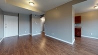This single-floor residence offers a cohesive and functional layout designed for comfortable everyday living. Throughout the space, rich dark hardwood-style flooring contrasts attractively with light gray walls and crisp white baseboards, creating a clean, modern aesthetic.

The open living area is generously proportioned, providing ample room for a variety of furniture arrangements. A tray ceiling with a ceiling fan adds architectural interest and dimension to the space, while a sliding glass door leads directly to a private balcony, filling the room with natural light and offering outdoor access.

The galley-style kitchen is efficiently designed with rich cherry-toned cabinetry lining both sides, complemented by granite-style countertops and a double stainless steel sink. Black appliances, including a refrigerator and dishwasher, complete the kitchen's cohesive look. A separate interior door in the kitchen provides additional storage or utility access.

The bathroom is well-appointed with a full tub-and-shower combination featuring white tile surround, a cherry wood vanity with a granite-style countertop, and coordinating hardwood-style flooring that ties the space together. The consistent use of gray tones, warm wood cabinetry, and durable flooring throughout gives this residence a unified, move-in-ready character.
