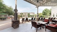 This expansive outdoor amenity space is designed to deliver a resort-like experience, centered around a generously sized freeform swimming pool with a smooth concrete surround and built-in entry steps with stainless steel handrails. Natural stone rockwork accents frame portions of the pool's edge, adding an organic, upscale aesthetic. Surrounding the pool, a wide sundeck accommodates numerous chaise lounges and shade umbrellas, providing abundant space for relaxation.

Adjacent to the pool, a substantial covered patio area features a white-painted pergola supported by stone-clad columns, offering shade while maintaining an open, airy atmosphere. Ceiling fans mounted within the pergola enhance comfort during warmer months. This shaded zone is furnished with wicker-style lounge chairs and dining sets with red cushions, suitable for both casual gatherings and outdoor dining.

A standout feature of the patio is a full-height stacked-stone outdoor fireplace, flanked by comfortable seating with green cushions, creating an inviting gathering spot during cooler evenings. The adjacent building facade showcases a refined architectural design with arched windows, stone and stucco exterior detailing, and a distinctive cupola roofline. Mature trees and landscaped greenery throughout the space provide a natural backdrop, enhancing the overall ambiance.