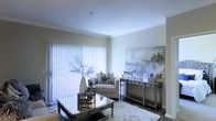 This single-floor space presents a thoughtfully designed living area finished in a neutral, light-toned palette that contributes to a bright and airy ambiance. Crown molding lines the ceiling, adding a refined architectural detail that elevates the overall aesthetic. A large sliding glass door fitted with vertical blinds serves as the room's primary light source, allowing generous amounts of natural light to filter through and illuminate the space.

The living area is furnished with a plush cream armchair accented by a patterned throw pillow, complemented by a mirrored console table that reflects light and enhances the sense of openness. A glass-topped coffee table with metallic legs anchors the seating area over a soft, textured area rug. Decorative elements include sculptural vases, a metallic figurine, and a large abstract canvas artwork in muted grey and yellow tones, lending the space a contemporary, gallery-like character. Dark hardwood-style flooring provides a grounding contrast to the lighter furnishings and walls, completing the polished, cohesive look of the interior.
