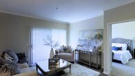 This single-floor space presents a thoughtfully designed living area finished in a neutral, light-toned palette that contributes to a bright and airy ambiance. Crown molding lines the ceiling, adding a refined architectural detail that elevates the overall aesthetic. A large sliding glass door fitted with vertical blinds serves as the room's primary light source, allowing generous amounts of natural light to filter through and illuminate the space.

The living area is furnished with a plush cream armchair accented by a patterned throw pillow, complemented by a mirrored console table that reflects light and enhances the sense of openness. A glass-topped coffee table with metallic legs anchors the seating area over a soft, textured area rug. Decorative elements include sculptural vases, a metallic figurine, and a large abstract canvas artwork in muted grey and yellow tones, lending the space a contemporary, gallery-like character. Dark hardwood-style flooring provides a grounding contrast to the lighter furnishings and walls, completing the polished, cohesive look of the interior.
