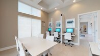 This single-floor business center is designed with a clean, contemporary aesthetic that promotes productivity and focus. The space is finished throughout in soft warm neutrals — taupe walls, crisp white trim, and large-format light tile flooring — creating a cohesive and professional atmosphere.

The main area features a row of three semi-private computer workstations, each separated by white divider panels and equipped with monitors. White ergonomic rolling chairs provide comfortable seating at each station, while framed abstract artwork displayed above the workstations adds a refined visual touch. A separate workstation area includes a printer cabinet and additional desk space, offering a dedicated zone for document handling.

A bright conference area accommodates a rectangular table surrounded by matching white chairs, ideal for small group meetings or collaborative work. Floor-to-ceiling roller shades filter abundant natural light through expansive windows, softening the glow while maintaining an open, airy feel. The coffered ceiling detail adds architectural interest and a sense of height to the room.

Throughout the space, recessed lighting ensures consistent illumination, complementing the natural light. The overall design balances functionality with a polished, modern sensibility, making it a well-equipped and inviting environment for business and professional use.