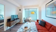 This well-designed one-bedroom, one-bathroom unit offers a thoughtfully laid-out single-level floor plan with modern finishes throughout. The kitchen serves as a central highlight, featuring high-gloss white cabinetry extending to the ceiling, white quartz countertops, a subway tile backsplash, and a full suite of stainless steel appliances including a French door refrigerator and built-in microwave. A spacious kitchen island with an undermount sink provides additional prep space and accommodates seating for multiple guests, making it ideal for casual dining and entertaining.

The open living area flows naturally from the kitchen, featuring warm hardwood-style flooring, recessed lighting, and a large window that fills the space with natural light and urban views. The bedroom continues the bright aesthetic with large floor-to-ceiling windows overlooking the city skyline, offering an airy and connected feel to the surrounding environment.

The bathroom is finished in a clean, contemporary style with neutral tile flooring, a walk-in glass-enclosed shower with full tile surround and rain showerhead, and a streamlined white vanity. The overall design is cohesive and modern, emphasizing clean lines, neutral tones, and functional use of space throughout.