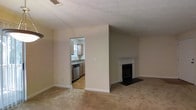 This single-floor residence offers a comfortable and functional layout designed for easy living. The main living area is finished with wall-to-wall carpeting in a warm neutral tone, creating a soft and inviting atmosphere. A classic fireplace with a white mantel and raised hearth serves as the room's focal point, adding character and a cozy element to the space. The textured ceiling adds a traditional touch throughout the living area. A wide doorway opening connects the living room to the kitchen, which features wood-toned cabinetry, a stainless steel dishwasher, a sink with a window above it allowing for natural light, and hard flooring. The kitchen's layout is efficient and well-organized, providing ample workspace. Neutral wall colors run consistently throughout both spaces, contributing to a clean and cohesive aesthetic. The open sightline between the living area and kitchen enhances the sense of flow and makes the overall space feel connected and functional. This home offers a straightforward, livable floor plan well-suited for everyday comfort.