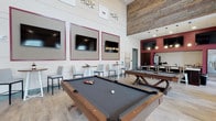This single-story recreational lounge is designed with entertainment and social interaction in mind. The space centers around two full-sized pool tables with dark felt surfaces and rich wood frames, positioned prominently on light wood-look flooring that runs throughout. Soaring double-height ceilings create an open, airy atmosphere, accentuated by a striking accent wall clad in reclaimed-style wood paneling that adds warmth and texture to the upper portion of the space.

The walls feature a clean white shiplap aesthetic with bold burgundy-framed flat-screen televisions mounted at comfortable viewing heights, complemented by decorative metallic wall accents above. Along one wall, high-top tables paired with modern gray bar stools provide seating for casual conversation or spectating. A rich red accent wall anchors a secondary lounge area in the background, illuminated by stylish pendant lighting that creates an intimate atmosphere within the larger open space.

A service or refreshment counter is visible in the background, suggesting amenities for guests. The overall design balances modern industrial touches with warm, inviting finishes, making it a well-appointed space suited for community gatherings and leisure activities.